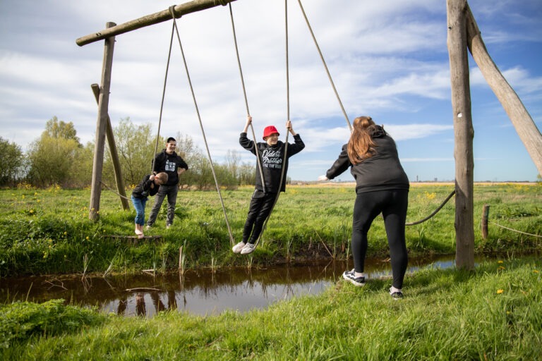 het hele gezin staat op de foto en de oudste zoon hangt in de touwen boven het water. moeder staat aan de overkant en vader en de middelste zoon staan nog aan de start om te kijken hoe die het doet