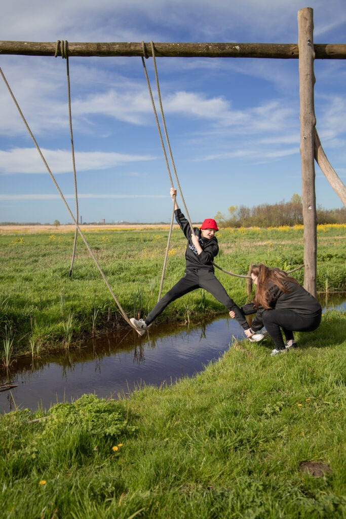 oudste zoon is in de touwen geklommen over de sloot heen en bereikt nu de overkant moeder pakt zijn voet om hem op het droge te trekken