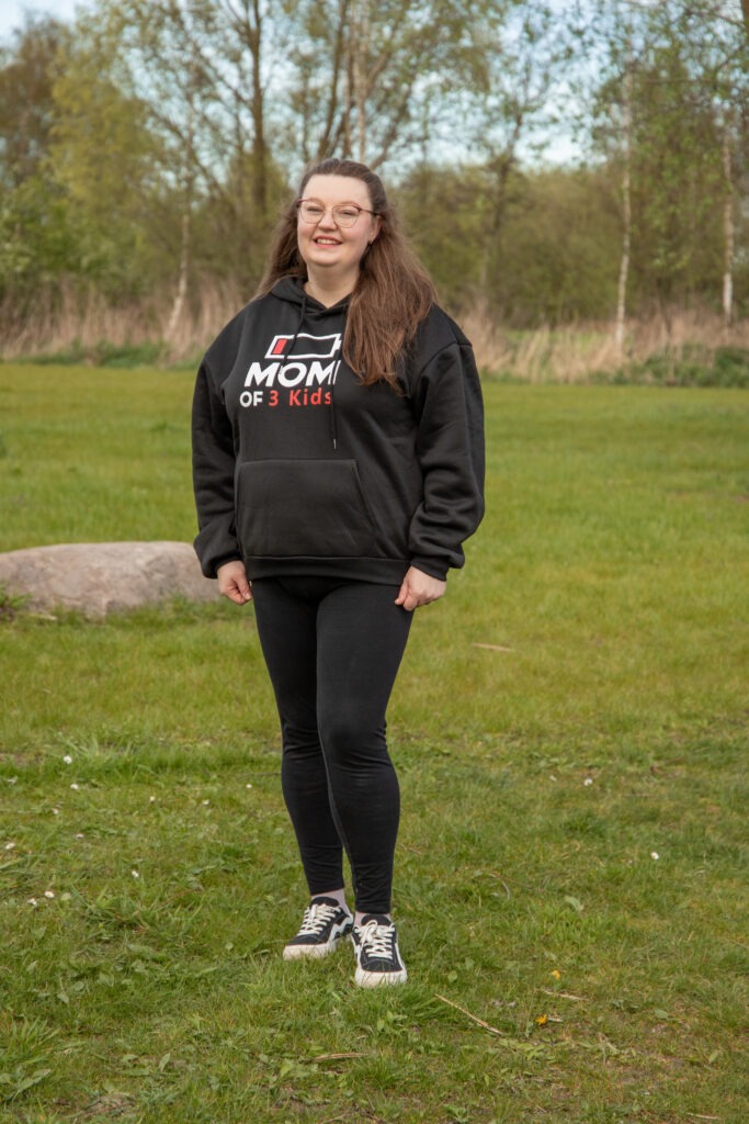moeder staat op de foto te lachen met een zwarte trui aan met daarop een teken van een lege batterij en met de tekst mom of 3 kids