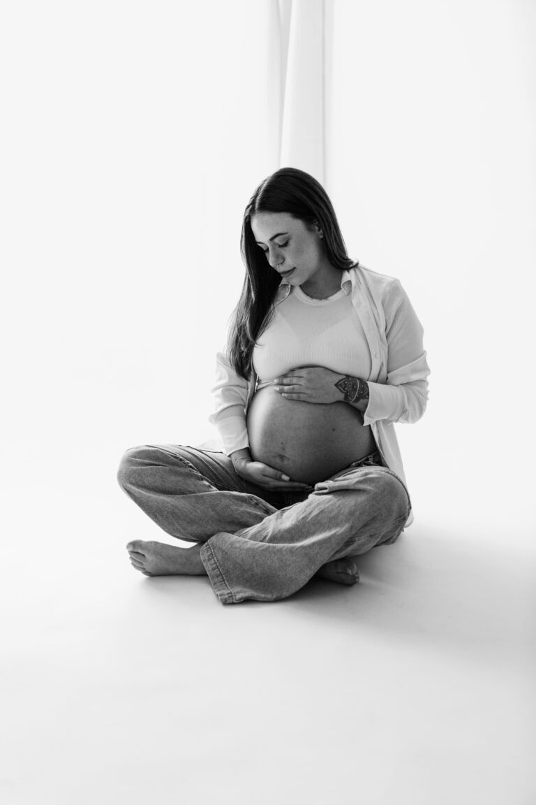 Zwangere vrouw zit in de daglichtstudio in kleermakerszit en houd haar buik vast en kijkt naar haar buik