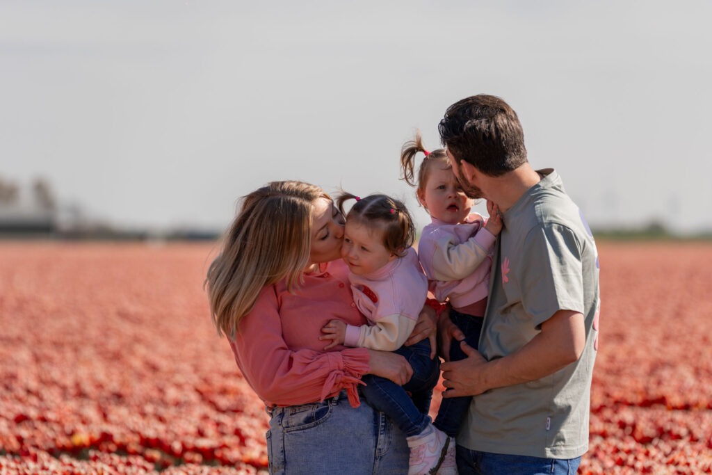 Knuffelmoment tijdens de familiefotoshoot in de tulpen – Moeder geeft haar dochter een kus, terwijl vader de andere dochter liefdevol vasthoudt tussen de bloemen.