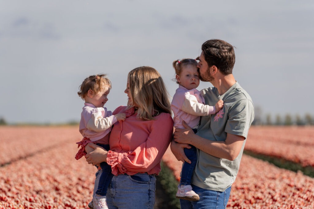 Familiekus in het tulpenveld – Vader geeft een kus aan zijn dochter, terwijl moeder en hun andere dochter elkaar liefdevol aankijken tussen de bloemen.