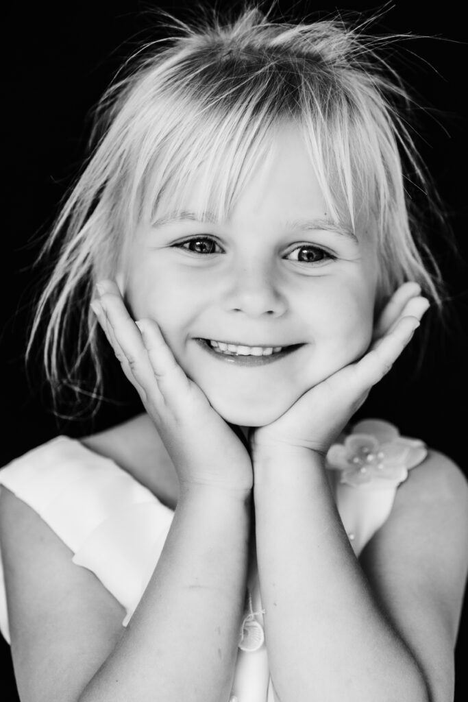 Blond meisje met een vrolijke glimlach, handen onder haar kin, voor een zwarte achtergrond – portret in zwart-wit.