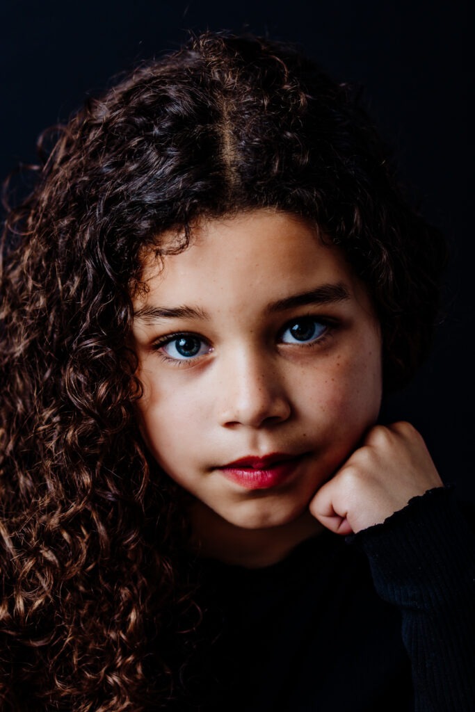 Meisje met krullend donker haar en blauwe ogen, kijkt rustig in de camera met haar hand onder haar kin, voor een zwarte achtergrond.
