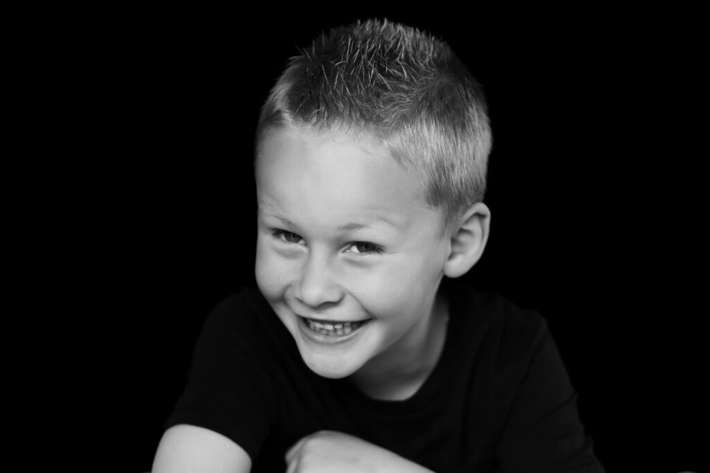 Lachende jongen met kort blond haar in een zwart shirt, leunend op zijn armen, vastgelegd in zwart-wit tegen een zwarte achtergrond.