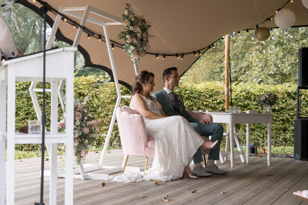 Bruid en bruidegom luisteren ontspannen naar de ceremonie Het bruidspaar zit samen op een zachtroze stoeltje onder een stretchtent, luisterend naar een spreker tijdens de buitenbruiloft.