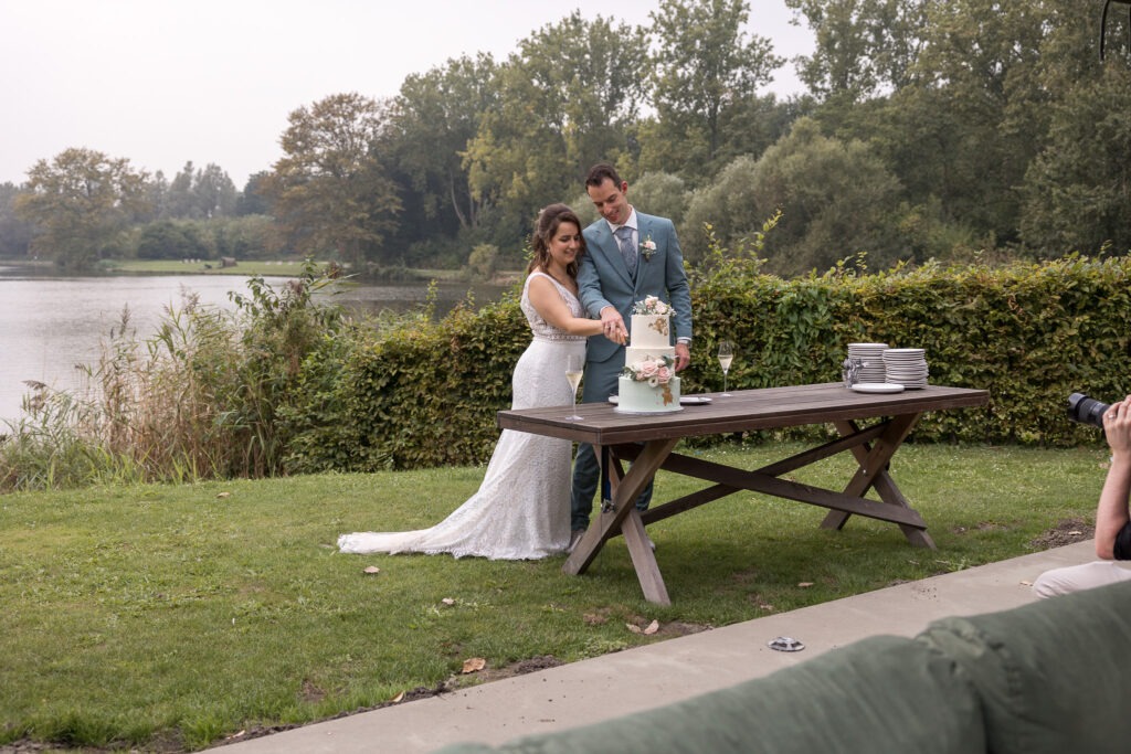 Bruidspaar snijdt samen de bruidstaart aan met uitzicht op het water Met uitzicht over een vijver snijdt het bruidspaar hun sierlijke witte bruidstaart aan, met champagneglazen op tafel en de fotograaf dichtbij.