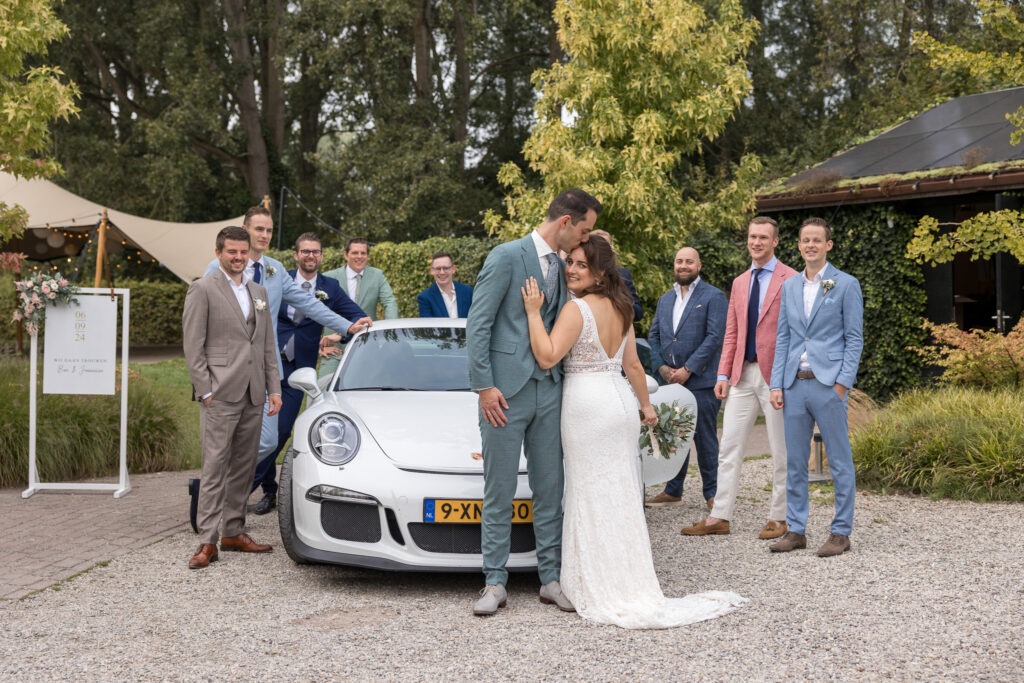 Bruidspaar poseert voor een witte Porsche met vrienden Het bruidspaar staat knus voor een sportauto, omringd door mannelijke gasten in kleurrijke pakken en glimlachen voor de groepsfoto.
