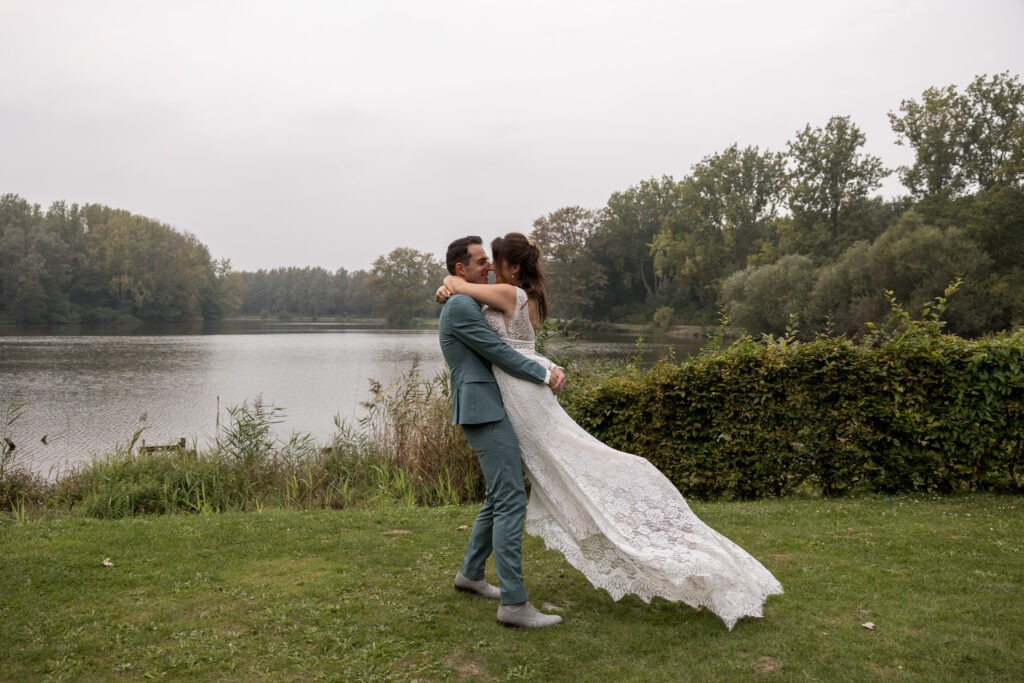 Bruidspaar bij het water, gedragen in een liefdevolle omhelzing De bruidegom tilt zijn bruid op aan de oever van het meer, met haar jurk die sierlijk achter haar wappert – een romantisch moment in de natuur.