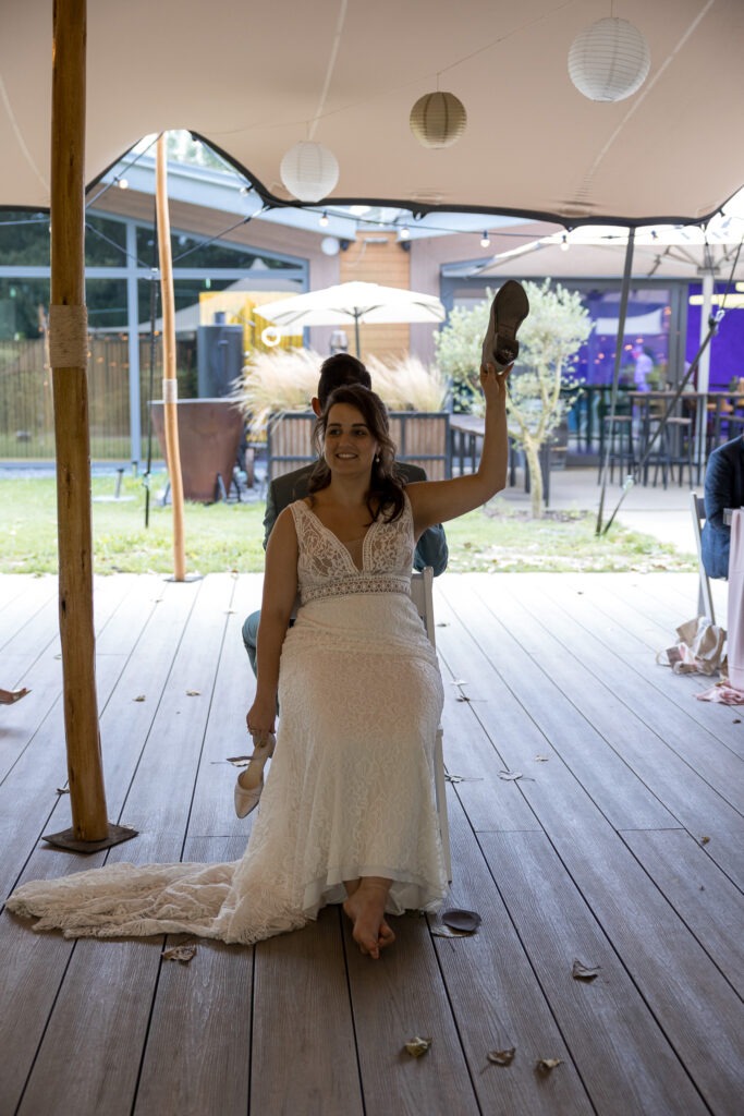 Het bekende schoenenspel tijdens het trouwfeest De bruid en bruidegom zitten rug aan rug tijdens het populaire schoenenspel, met een grote glimlach en schoenen in de lucht.
