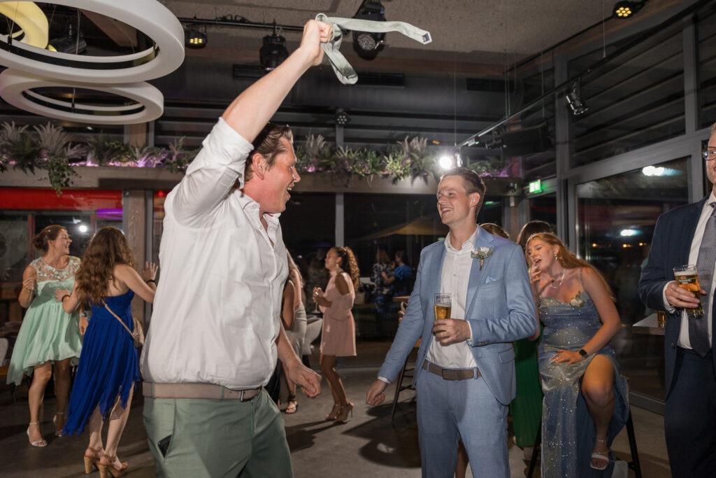 Feestganger zwaait met stropdas op de dansvloer Een gast danst uitbundig met zijn stropdas in de lucht, terwijl de bruidegom toekijkt met een glimlach en een biertje in de hand.