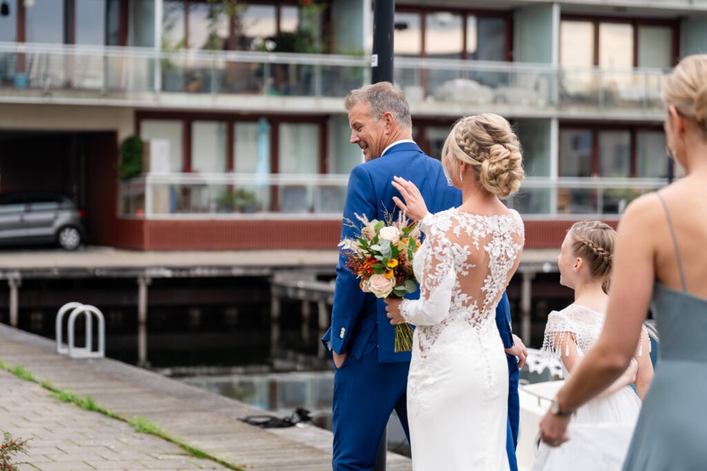 De bruid tikt de bruidegom op de schouder tijdens de first look De bruid nadert de bruidegom van achteren, haar hand op zijn schouder, met haar prachtige boeket en kanten jurk – het moment van de onthulling.
