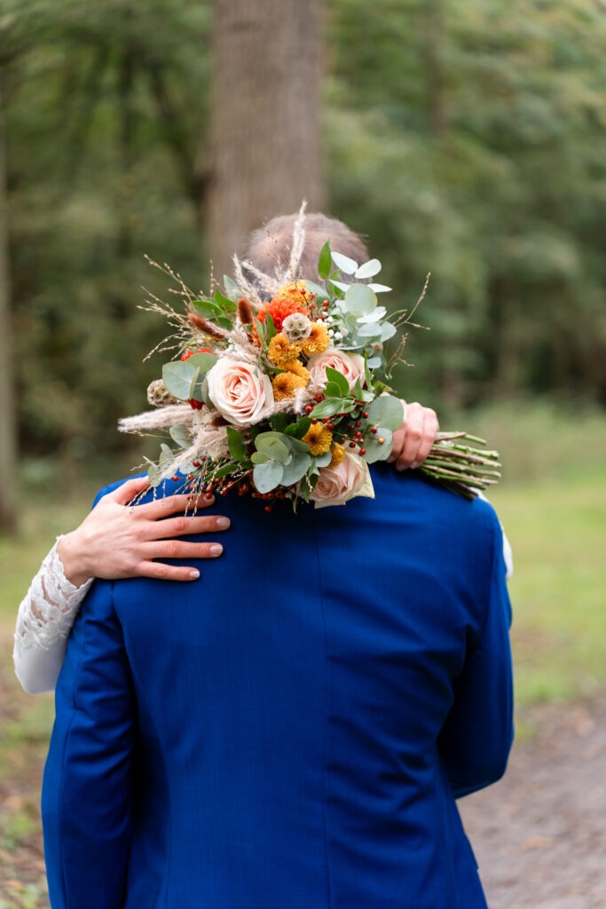 Detail van bruidsboeket achter de rug van de bruidegom Close-up van het kleurrijke bruidsboeket met rozen, eucalyptus en oranje accenten, vastgehouden achter de rug van de bruidegom.