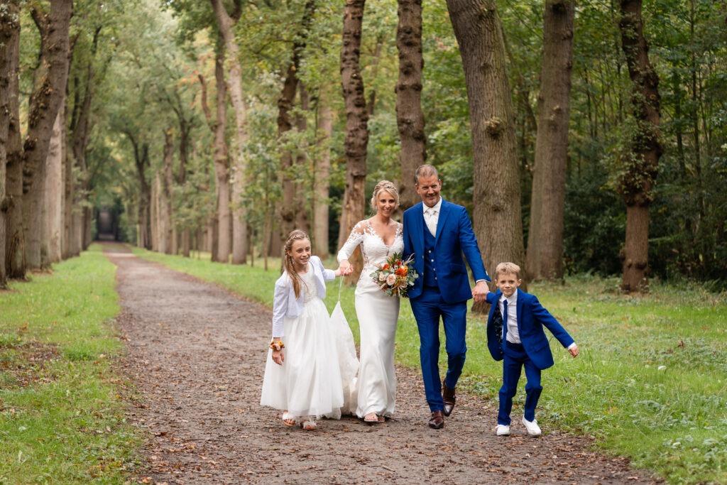 Bruidspaar loopt hand in hand met hun kinderen door het bos Het bruidspaar wandelt samen met hun dochter en zoon over een bospad, lachend en ontspannen tijdens hun trouwshoot.