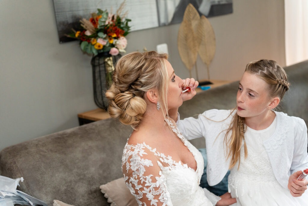 Dochter brengt lippenstift aan bij haar moeder, de bruid Een ontroerend moment thuis: de dochter helpt haar moeder met lippenstift terwijl de bruid rustig blijft zitten met haar boeket op de achtergrond.