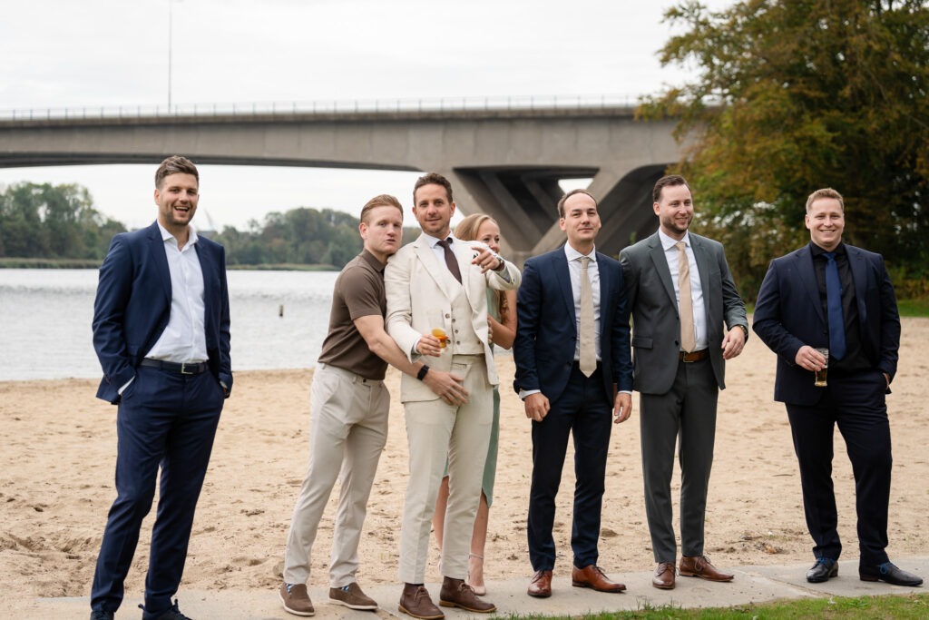Vriendengroep poseert bij het water tijdens de bruiloft Een groep vrienden in pak poseert lachend bij het strand en de brug op de achtergrond, met drankjes in de hand.