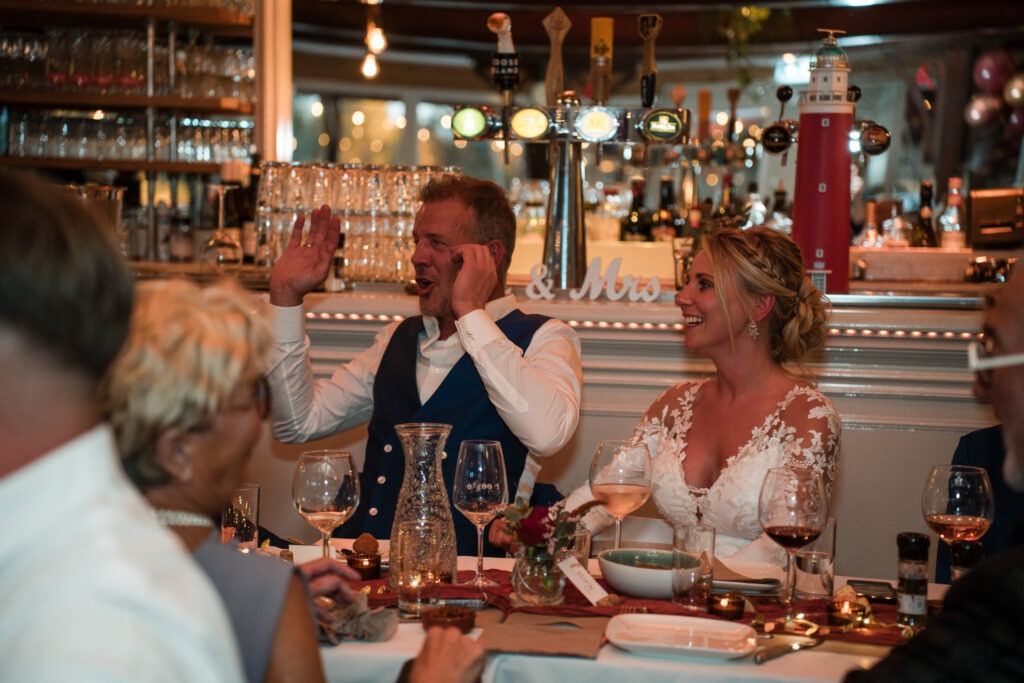 Bruidspaar lacht tijdens het diner bij de bruiloft De bruidegom gebaart enthousiast terwijl de bruid lachend toekijkt aan de dinertafel, omringd door familie en vrienden in een sfeervol verlichte zaal.