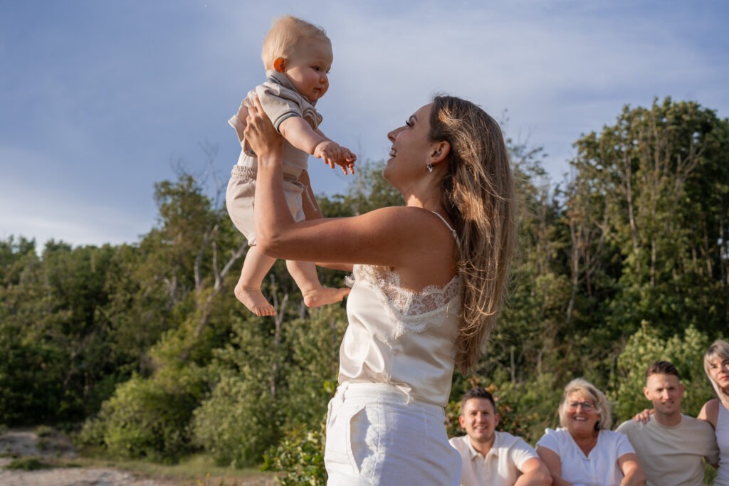 Moeder tilt haar baby lachend op in het avondlicht Tijdens de familieshoot in Rockanje tilt mama haar zoontje hoog in de lucht – beiden stralen, met familie op de achtergrond en zacht avondlicht over de duinen.