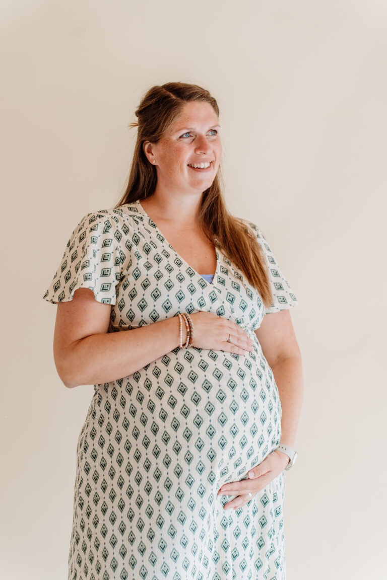 Zwangere vrouw lacht terwijl ze met haar handen haar buik vasthoudt, lichte achtergrond, zachte belichting.