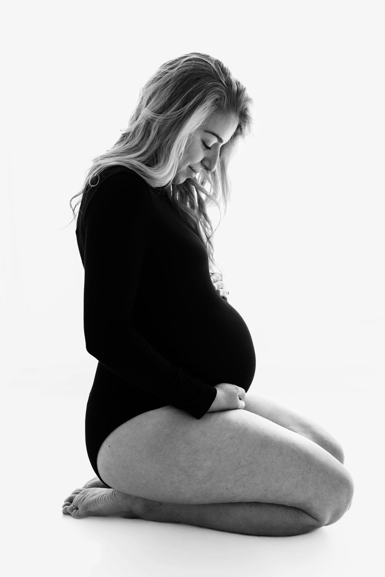 Zwangere vrouw die knielend poseert tijdens een zwangerschapsfotoshoot, met een zachte blik naar haar buik in een lichte studio.