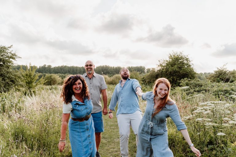 Vrolijk gezin dat hand in hand door het hoge gras wandelt tijdens hun familieshoot in de Broekpolder in Vlaardingen.