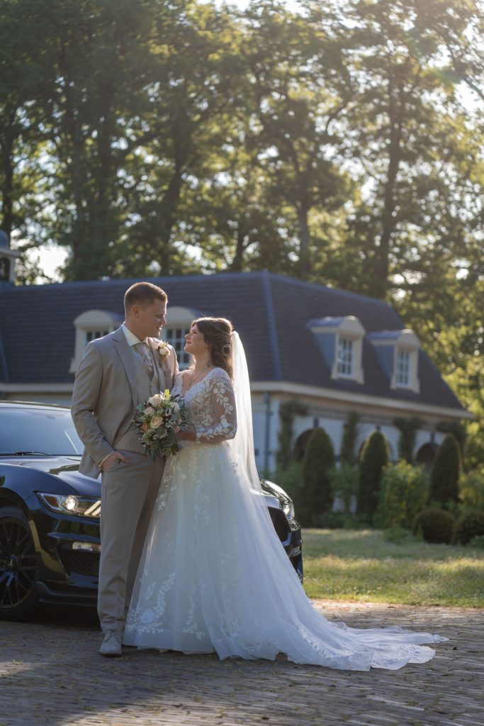 Pasgetrouwd bruidspaar poseert liefdevol bij een auto in een groene omgeving