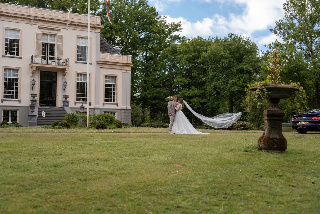 Bruid en bruidegom staan hand in hand op het gras voor een statig landhuis tijdens hun trouwdag