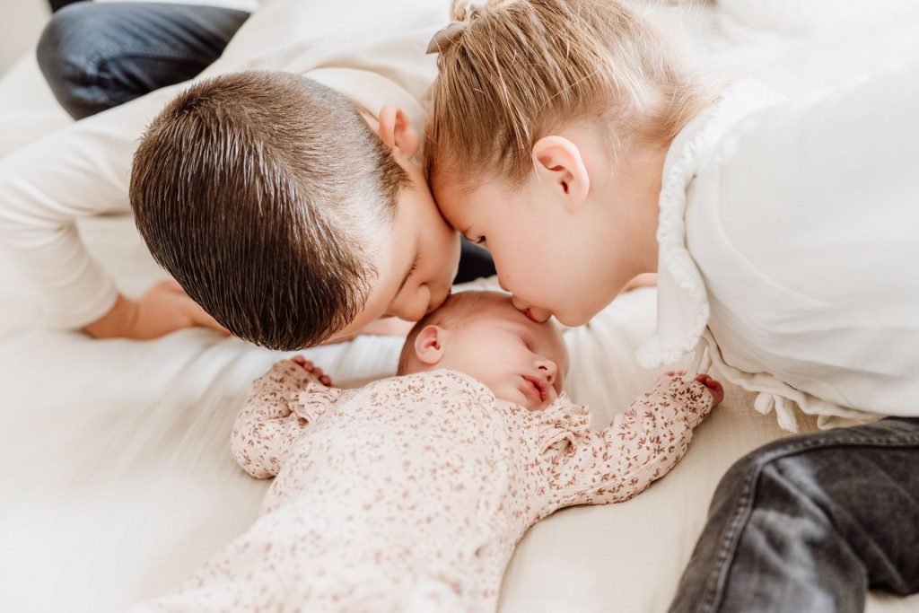Grote broer en zus geven allebei een kusje op het hoofd van pasgeboren Yara-Nove tijdens een newborn fotoshoot thuis in Zuidland.