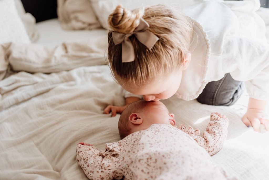 Grote zus buigt voorover en geeft een lief kusje op het hoofd van slapende newborn Yara-Nove, liggend op een linnen deken tijdens de fotoshoot thuis in Zuidland.