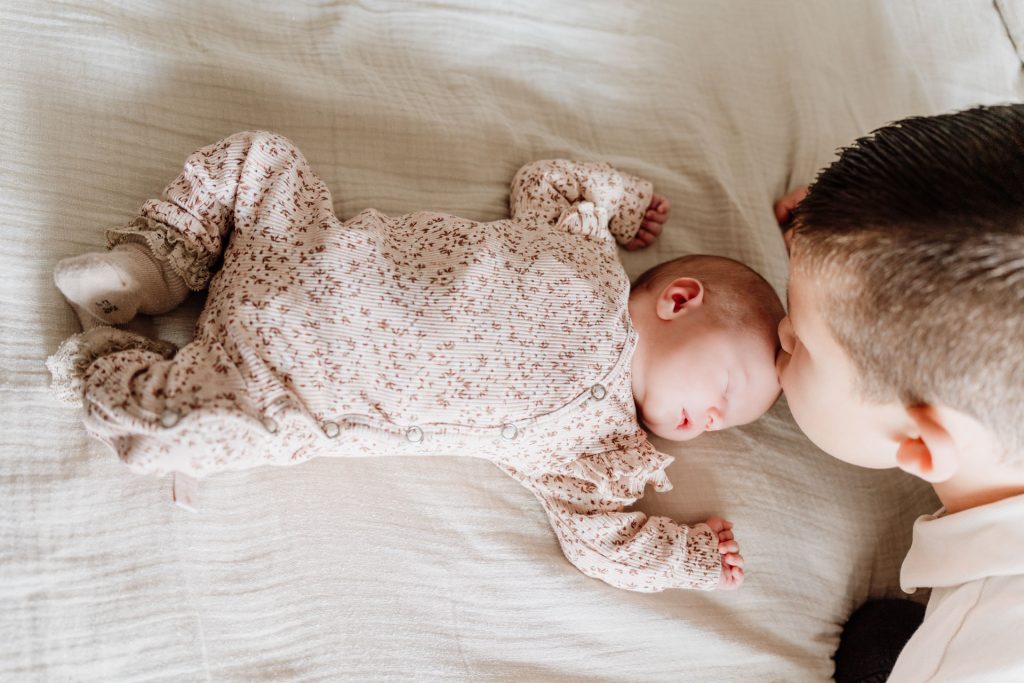Grote broer ligt naast slapende newborn Yara-Nove en kijkt vol liefde naar haar, vastgelegd tijdens een newborn fotoshoot thuis in Zuidland.