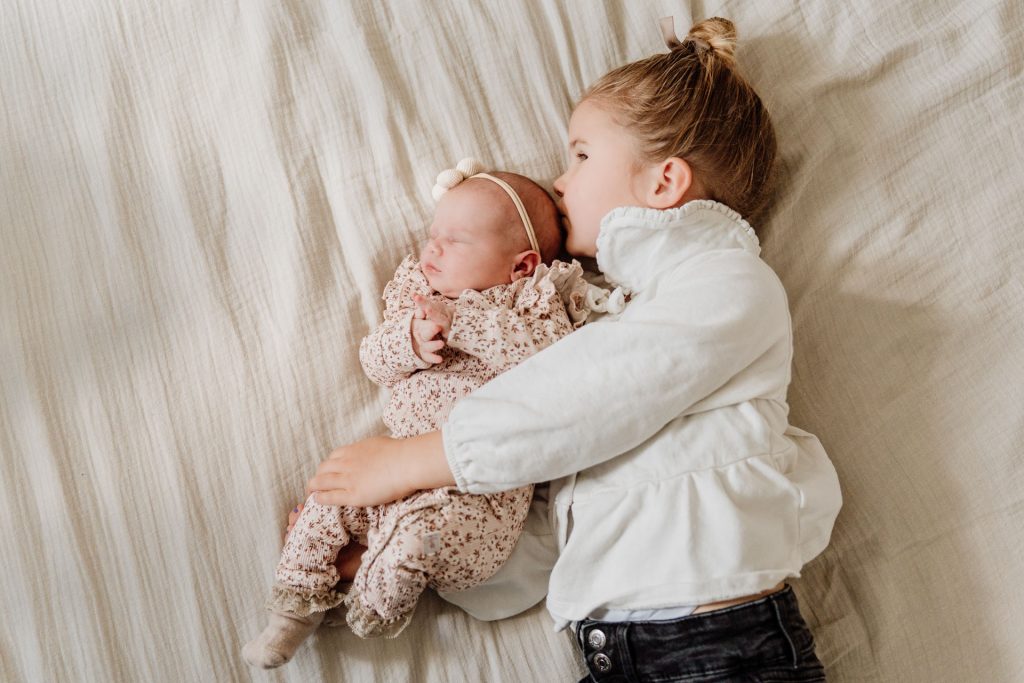 Grote zus houdt newborn Yara-Nove voorzichtig vast en ligt naast haar op bed, een intiem zusjesmoment tijdens de newborn fotoshoot thuis in Zuidland.