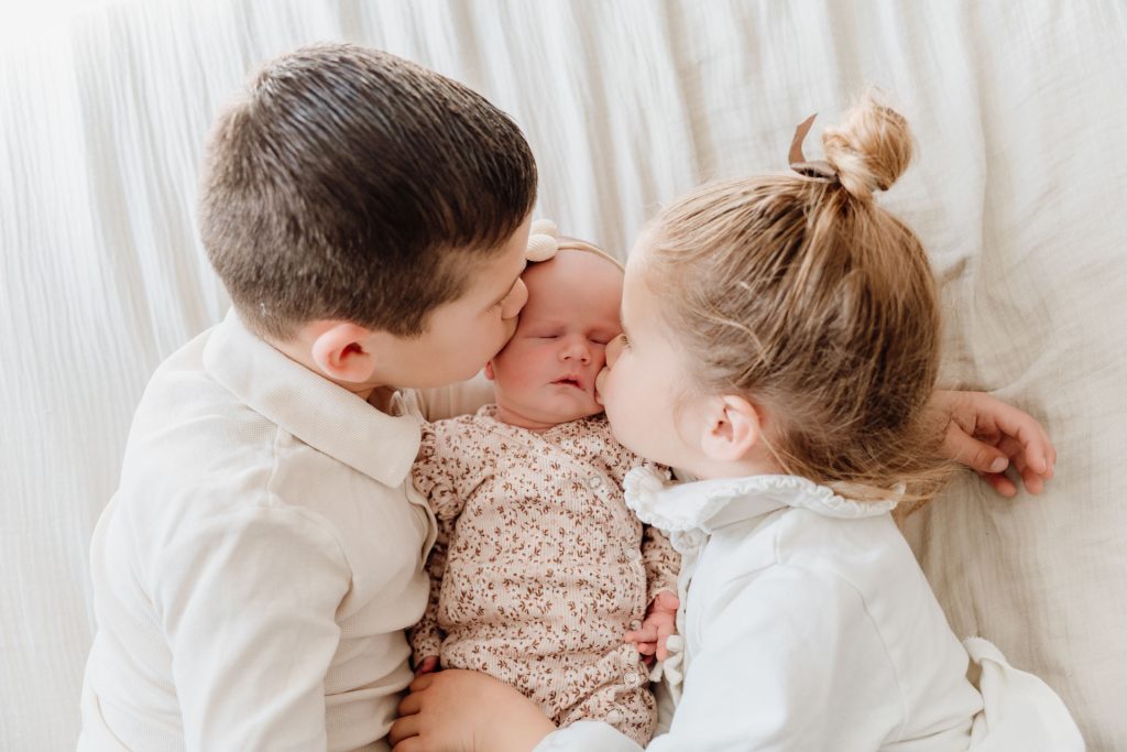 Broer en zus geven tegelijk een kusje op de wangetjes van slapende newborn Yara-Nove, vastgelegd tijdens een warme newborn fotoshoot thuis in Zuidland.