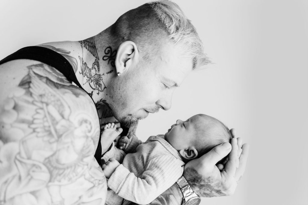 Mike houdt baby Ziva dicht tegen zich aan tijdens de newbornshoot in de studio, zijn tatoeages en het piepkleine kindje vormen een prachtig contrast
