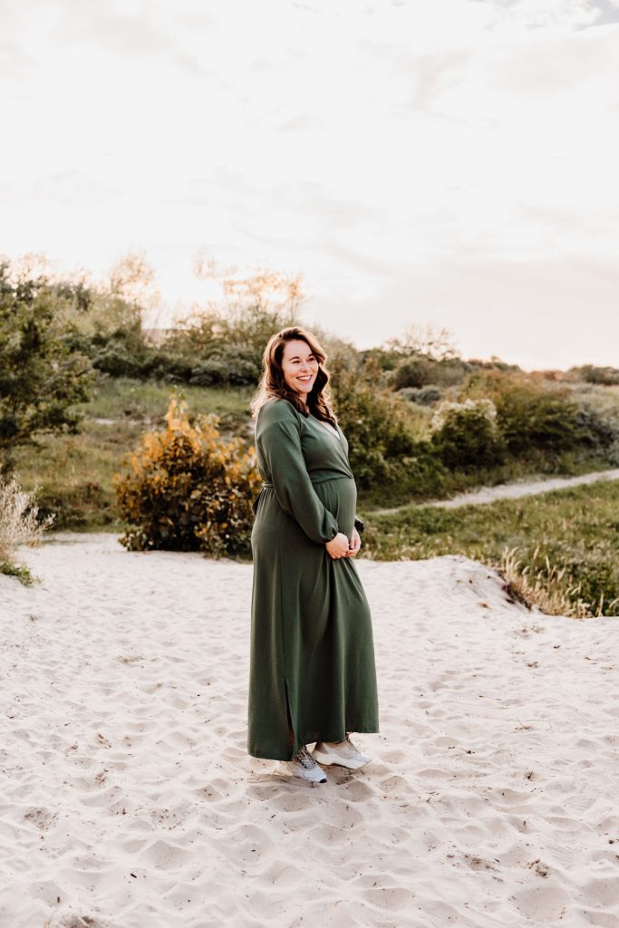 Zwangere vrouw in groene maxi-jurk staat lachend in de duinen van Rockanje en kijkt over haar schouder, zonsondergangssfeer