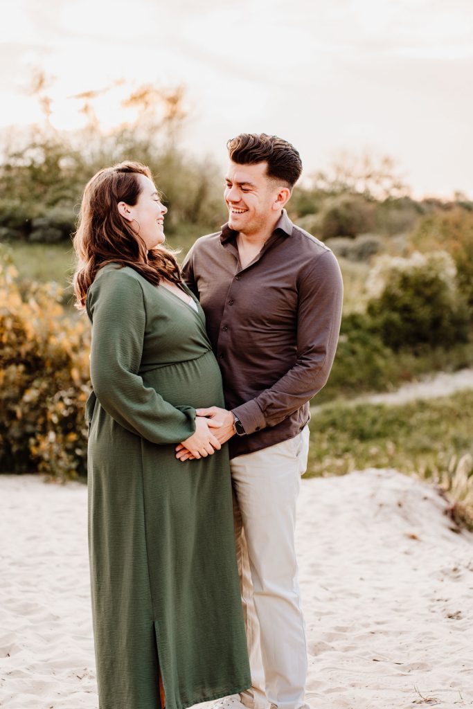 Zwangerschapsshoot met partner op strand Rockanje — stel lacht lachend naar elkaar met handen op de zwangere buik