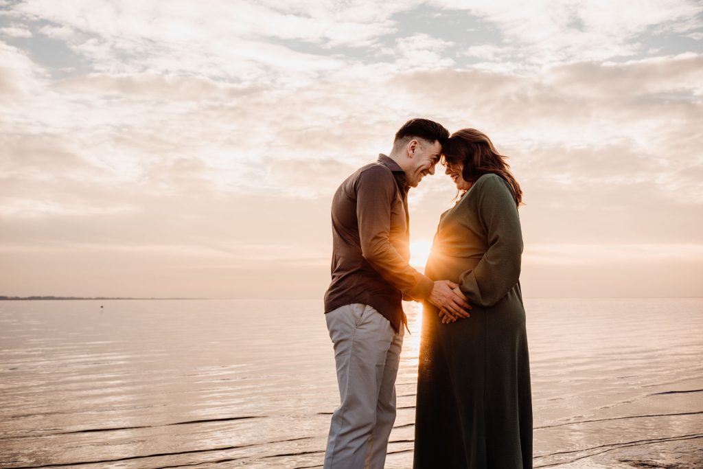 Zwanger stel staat hoofd tegen hoofd met de zon die precies tussen hen door schijnt tijdens zwangerschapsshoot strand Rockanje