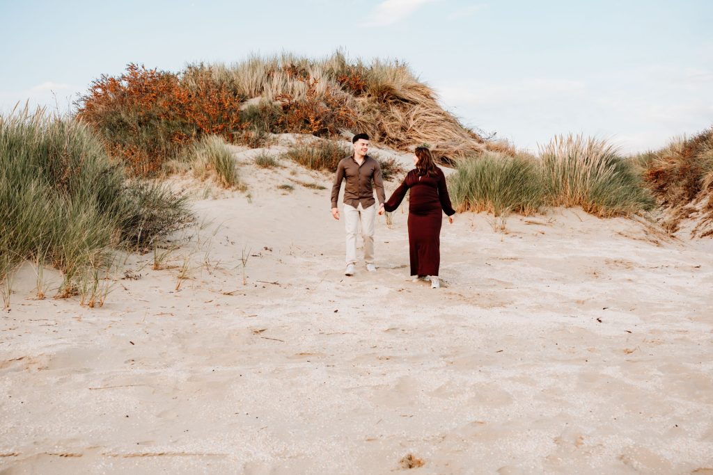 Stel wandelt hand in hand door de duinen van Rockanje tijdens zwangerschapsshoot strand Rockanje, bordeauxrode jurk, herfstsfeer