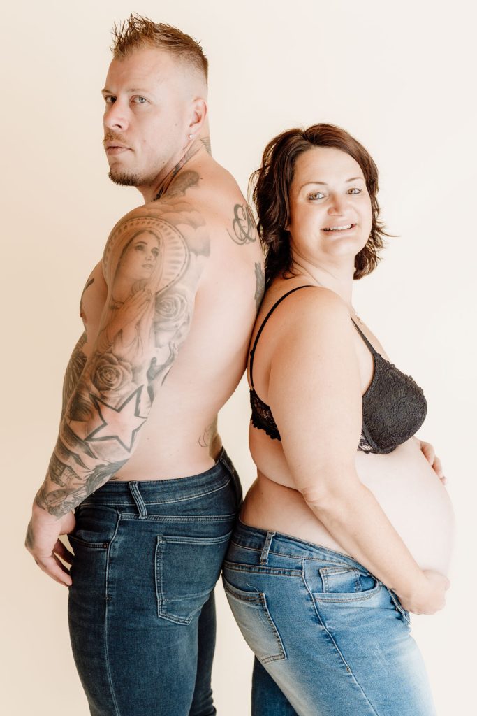Sanne en Mike staan rug aan rug in de studio, Mike kijkt stoer opzij terwijl Sanne glimlachend haar zwangere buik aanraakt