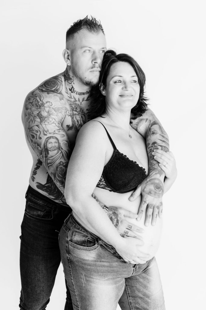 Zwart-wit foto van Sanne en Mike tijdens hun zwangerschapsshoot in de studio in Zuidland, Mike omarmt Sanne van achteren met zijn tatoeagerijke armen