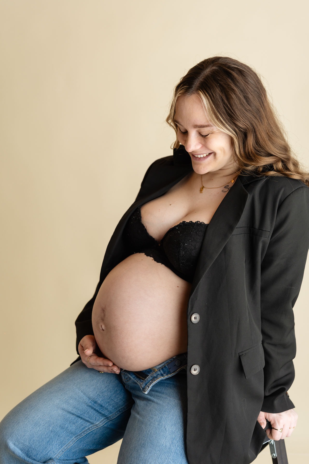 Zwangere vrouw in zwarte blazer lacht naar haar blote babybuik tijdens zwangerschapsshoot in de studio in Zuidland