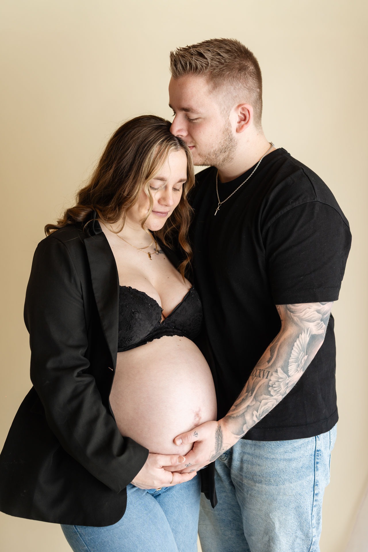 Partner kust zwangere vrouw op haar voorhoofd terwijl ze samen de babybuik vasthouden, zwangerschapsshoot Zuidland