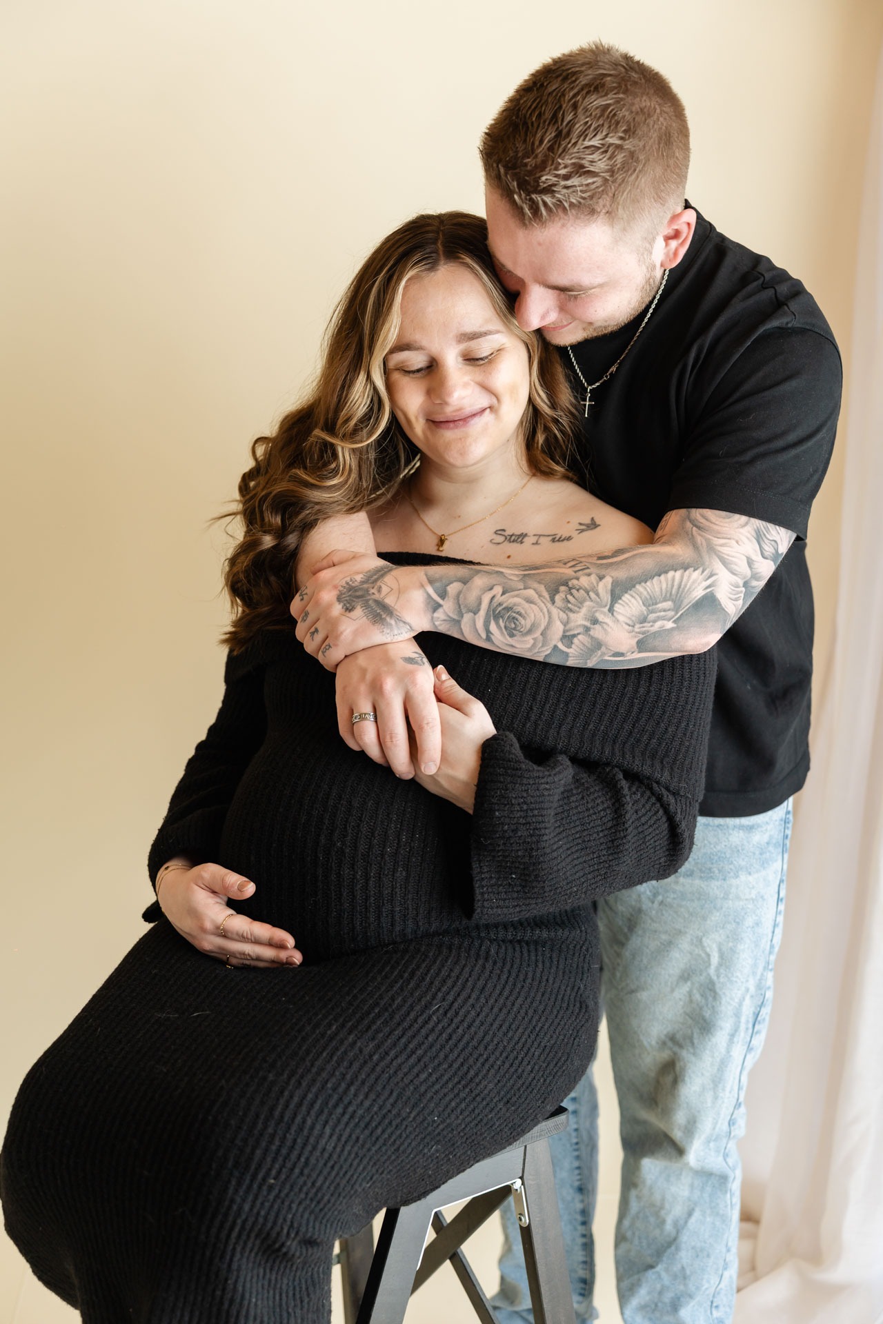 Zwanger stel omhelst elkaar liefdevol tijdens zwangerschapsshoot in de studio, zij in zwarte gebreide jurk