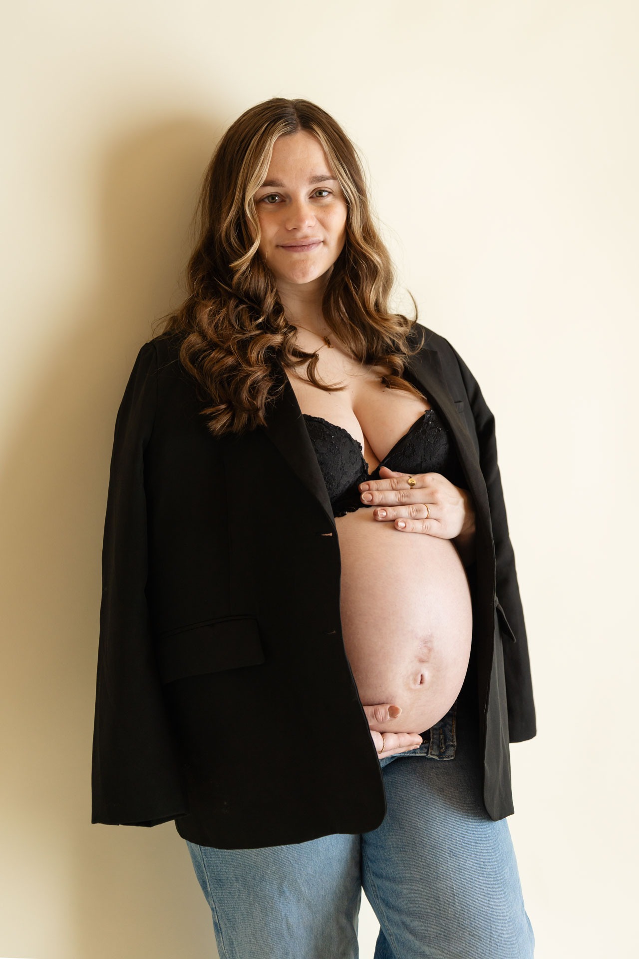 Zwangere vrouw in zwarte blazer en spijkerbroek toont haar babybuik tijdens zwangerschapsshoot bij Daphne's Fotografie