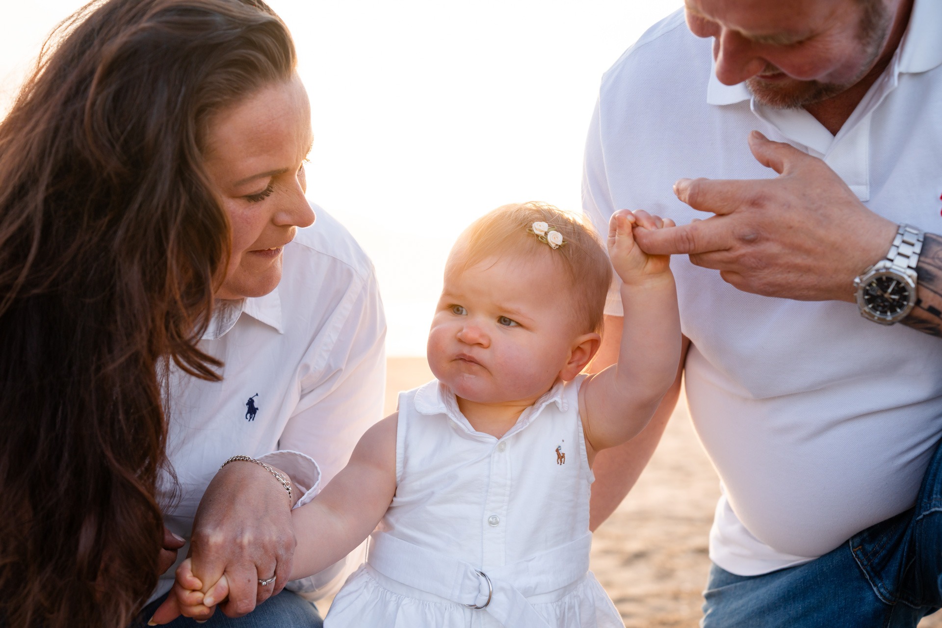 Meisje van één houdt papa's vinger vast terwijl beide ouders naar haar kijken tijdens familieshoot strand Rockanje